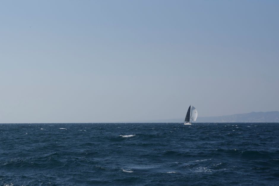 A lone sailboat glides through the open ocean with a clear blue sky horizon.