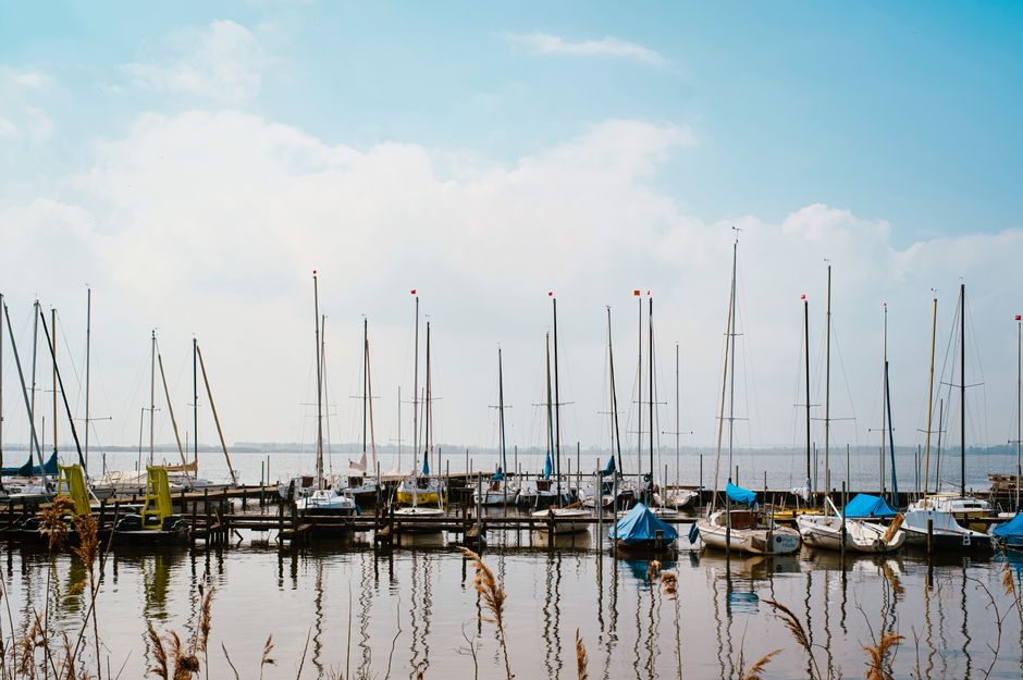 Peaceful harbor scene with sailboats moored under a bright blue sky, reflected on calm waters.